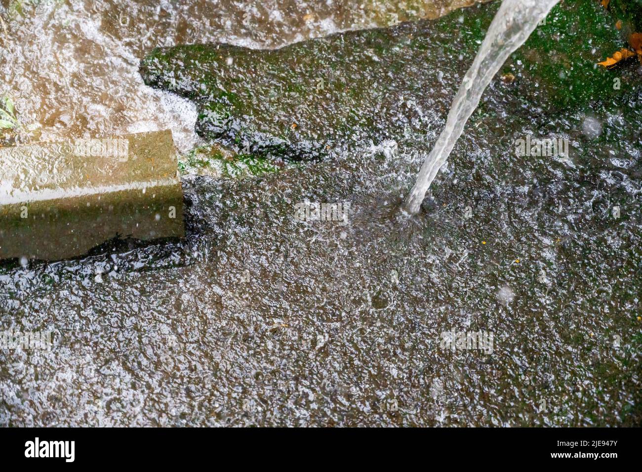 Verso il basso sulla strada della città. L'acqua piovana scorre lungo il marciapiede lungo il marciapiede. Il flusso denso di acqua cade dal tubo di drenaggio. Grandi gocce di pioggia. Estate th Foto Stock