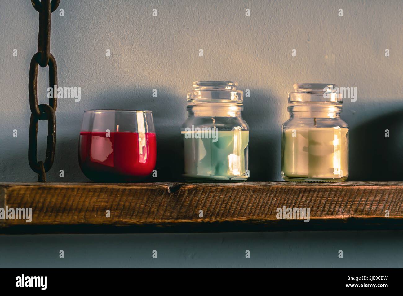 tre candele profumate colorate su un ripiano a parete rustico fatto di vecchio legno di fienile rigenerato con catena arrugginita Foto Stock