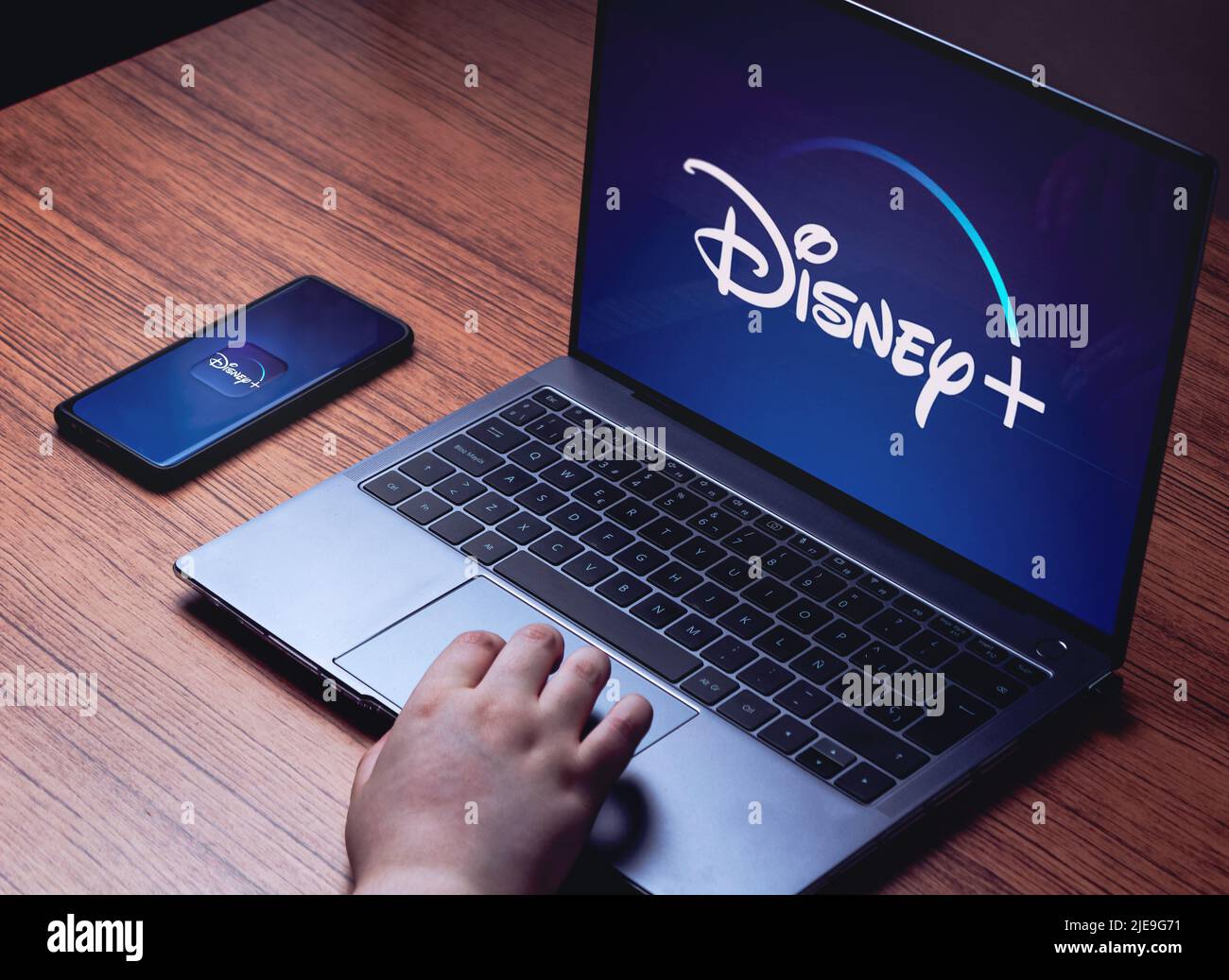 Logo Disney+ sugli schermi di notebook e smartphone. Scrivania in legno con laptop e smartphone in streaming Disney+. Foto Stock