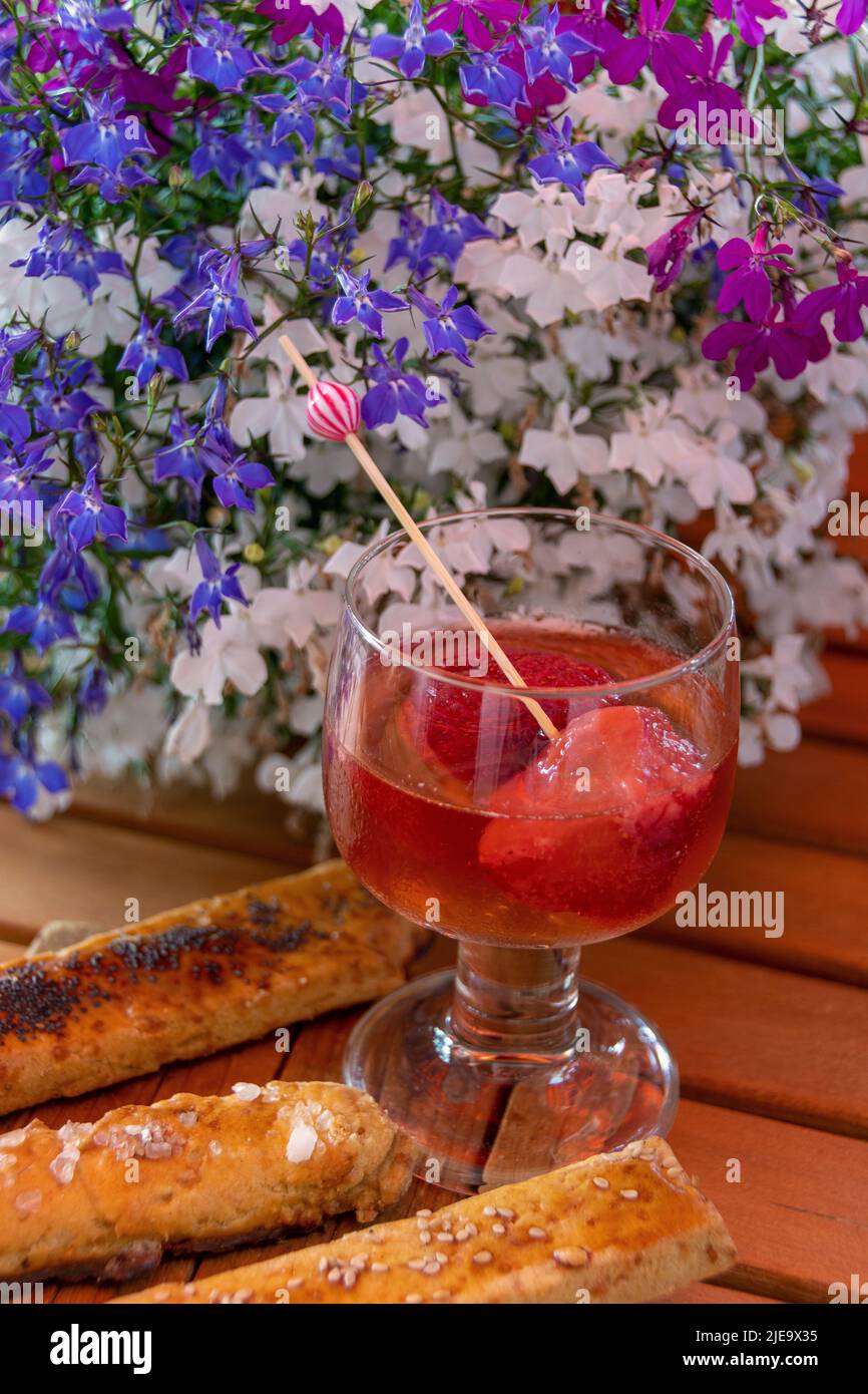 Im Hintergrund Blumen , im Mittelpunkt ein Glas Bowle mit Erdbeeren, zu Füssen des Glases liegen Gebäckstangen. Angerichtet ist auf einem Holztisch Foto Stock