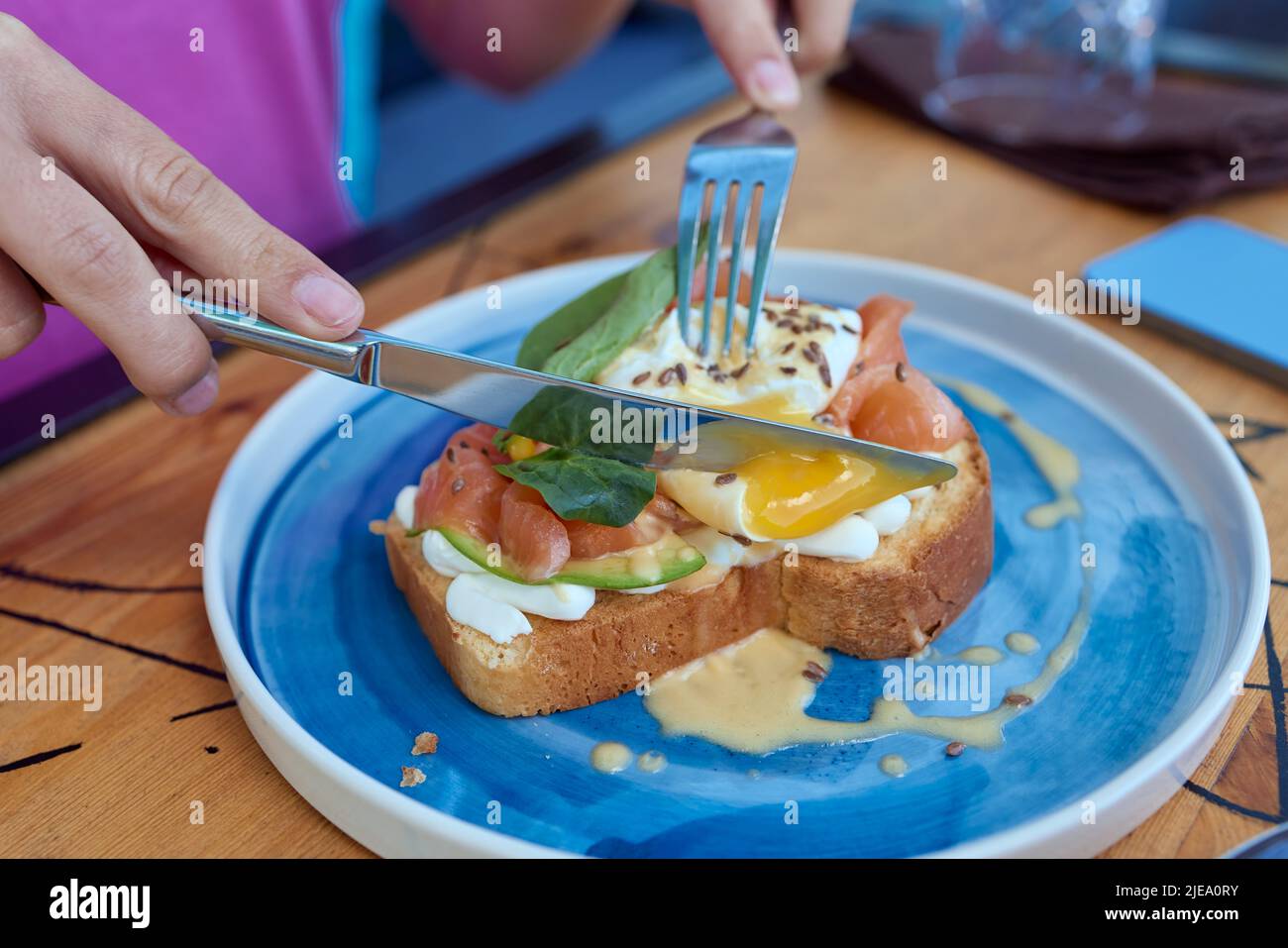 La giovane donna mangia la colazione con il coltello e la forchetta, la mattina in un caffè. Colazione sana con pane tostato integrale con avocado, uova in camicia con sala verde Foto Stock