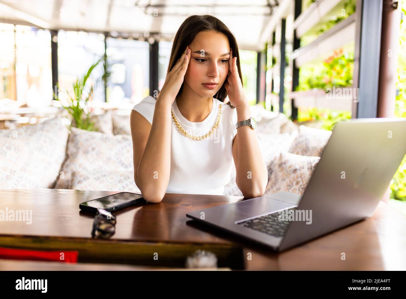 Donna stressata usando il laptop al caffè, seduto al tavolo con blocco note e tazza di caffè da solo, toccando i suoi templi, sperimentando burnout professionale, h Foto Stock