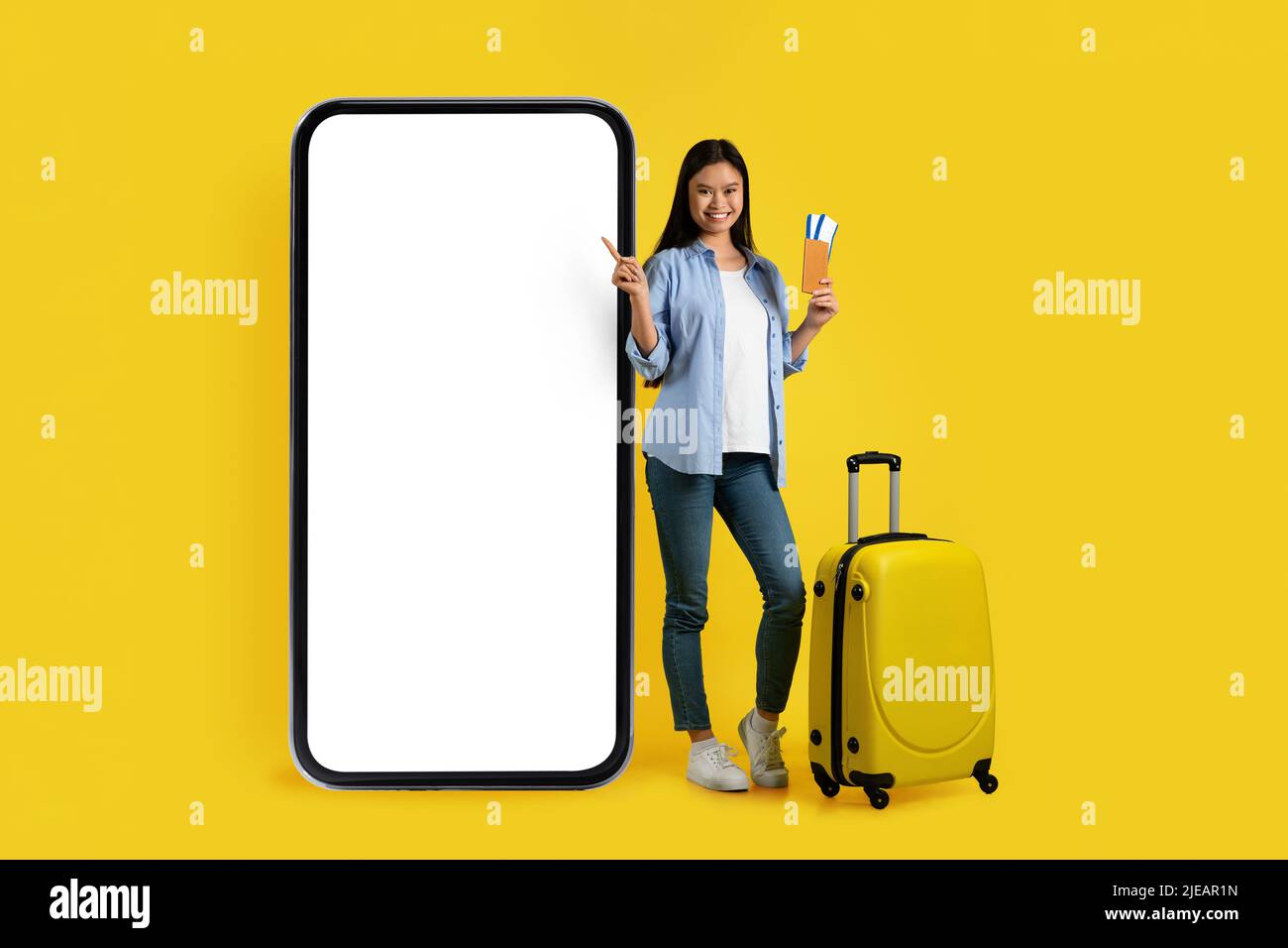 Felice giovane donna asiatica con valigia, passaporto e biglietto puntino dito su un enorme telefono con schermo vuoto Foto Stock