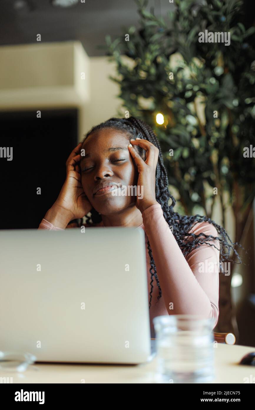 Depressa afroamericana donna che tiene le mani sulla testa con gli occhi chiusi e che lavora sul laptop in chioseup caffè casa. Ragazza nera che soffre di dolore Foto Stock