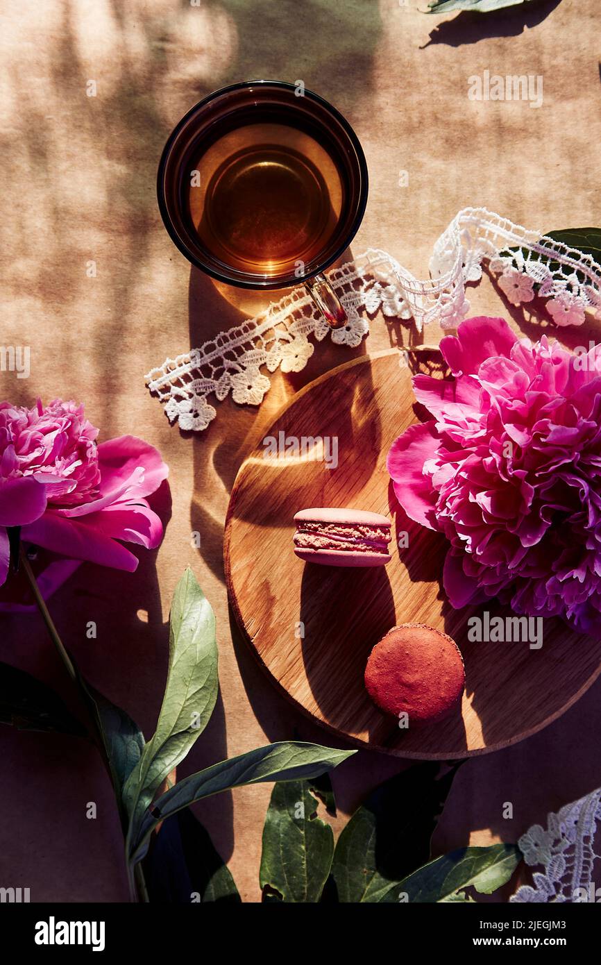 Dessert di macaron luminosi e tè alle erbe tra fiori rosa fioriti di peonia sotto le ombre dure alla moda nella terrazza. Estate estetica, relax, pensieroso, meditativo, stile di vita di buon umore Foto Stock