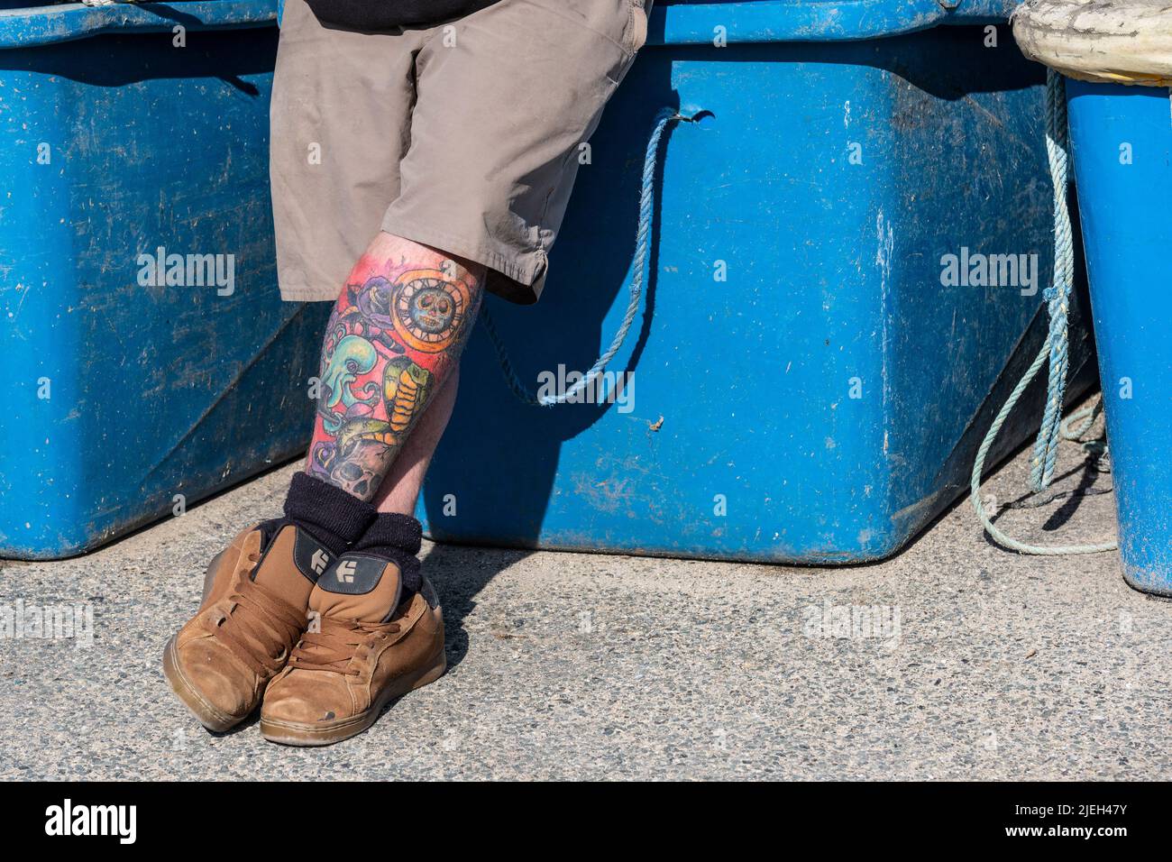 Un uomo che si rilassa contro i bidoni di stoccaggio con le gambe tatuate incrociate. Foto Stock