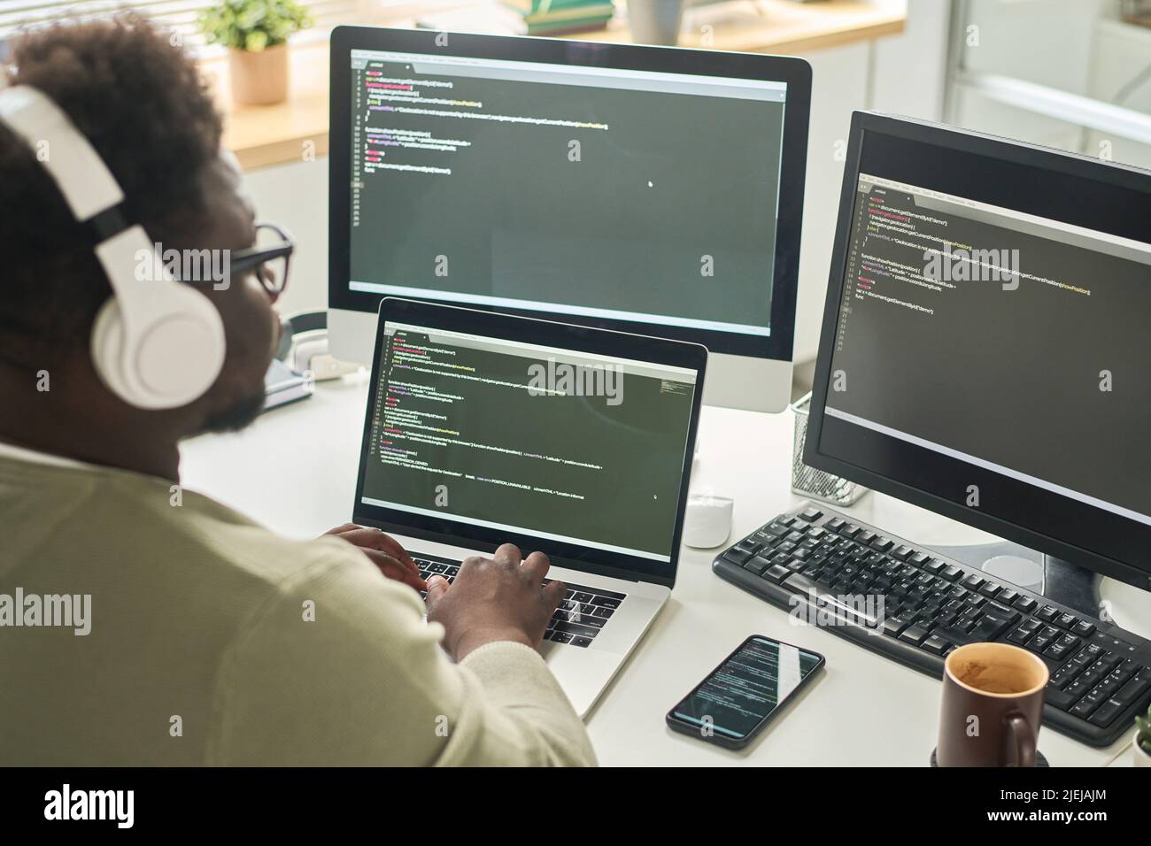 Vista posteriore di un uomo africano in cuffie wireless scrivere codici sul computer seduto sul posto di lavoro in ufficio IT Foto Stock