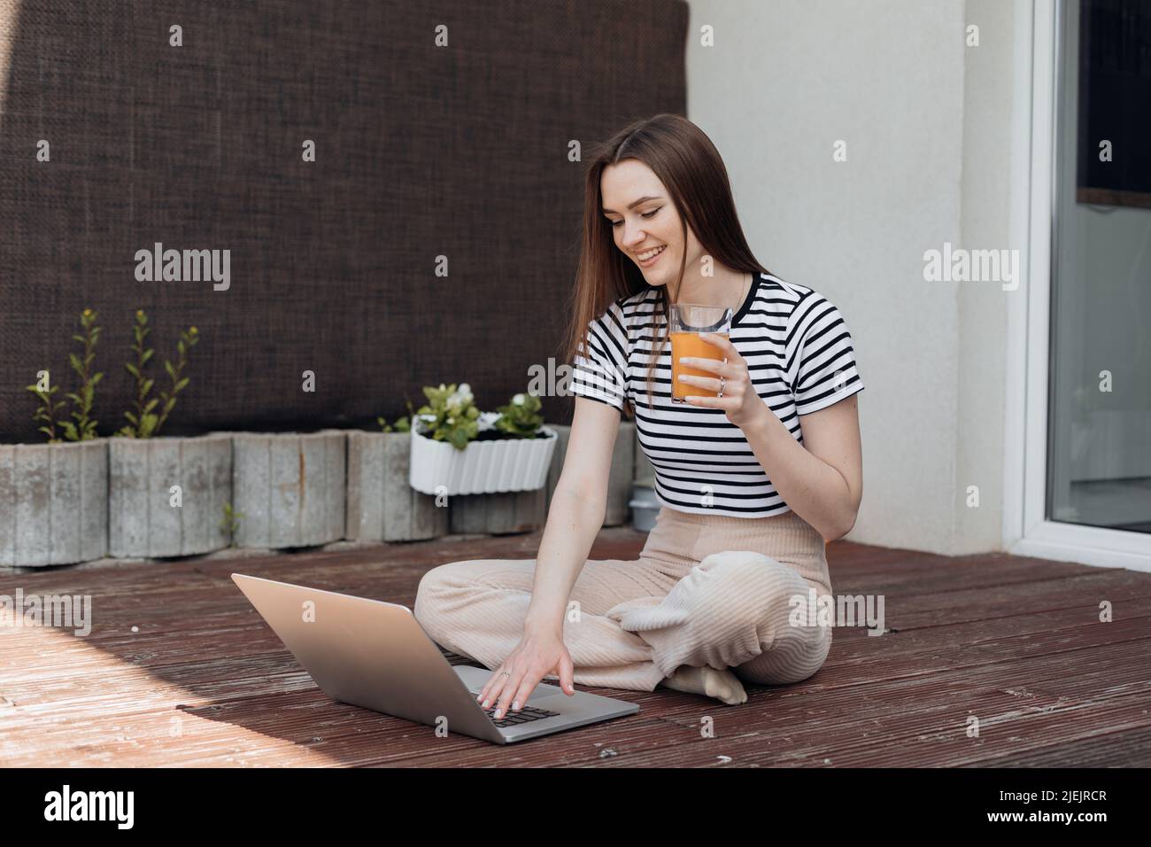 Sorridente giovane donna seduta all'aperto in terrazza, lavorare in remoto online su un laptop e bere fresco succo vista laterale. Freelance, lavoro a distanza, affari Foto Stock