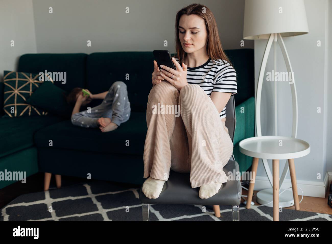 Donna bambino seduto su sedia e giocare al telefono, ragazzino sdraiato sul grande divano verde in soggiorno. Indifferenza verso il bambino, l'idlezza, il lavoro difettoso Foto Stock