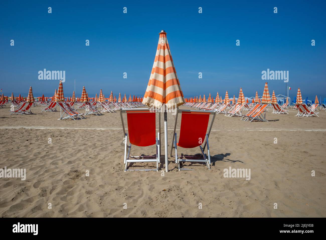 Una vista di sdraio rosse sulla spiaggia con ombrelloni arancioni e bianchi in una giornata di sole con un cielo azzurro. Foto Stock