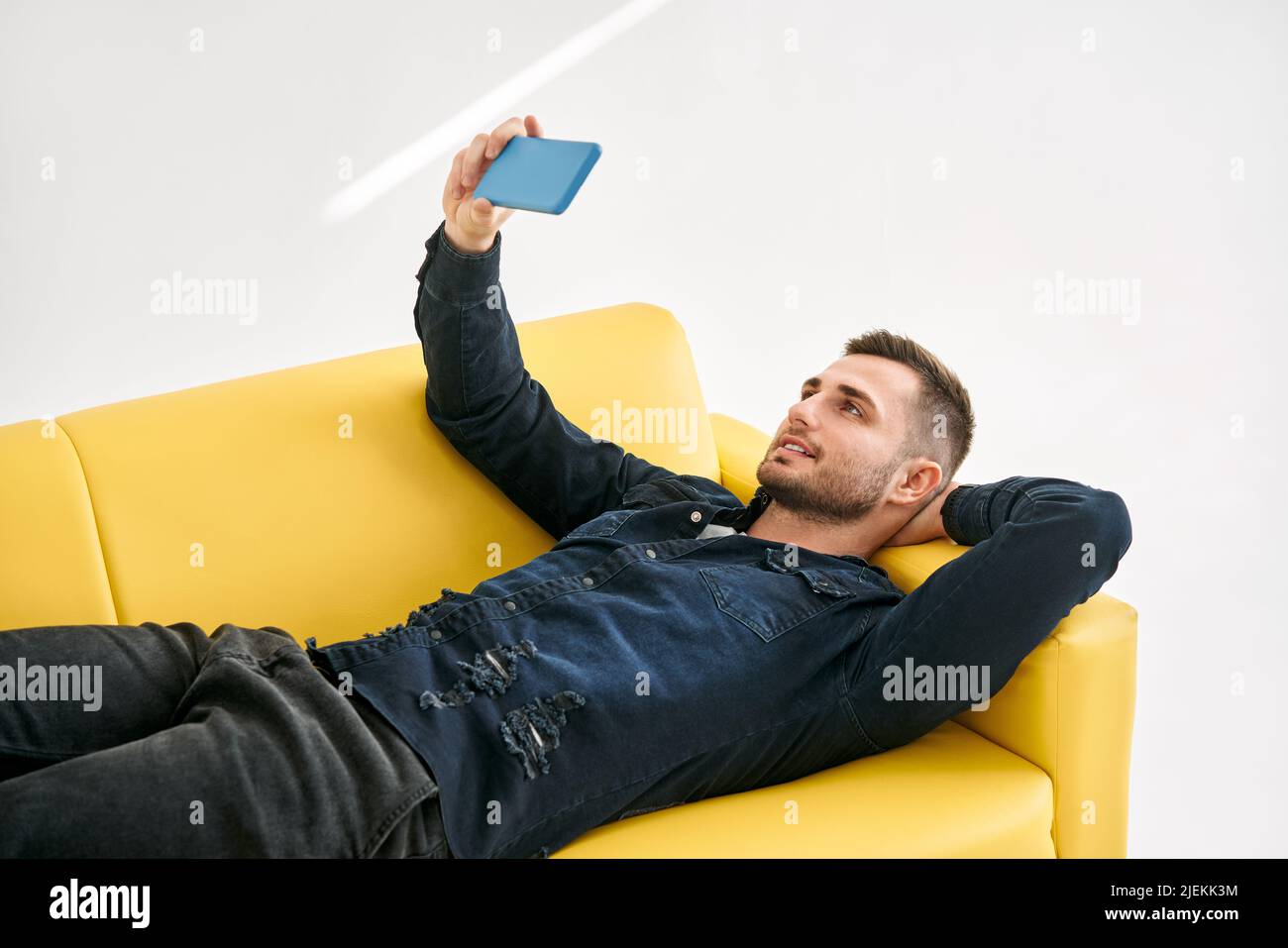 Uomo rilassato sdraiato sul divano giallo parlare in videochiamata dal suo cellulare su sfondo bianco. Concetto di social media Foto Stock