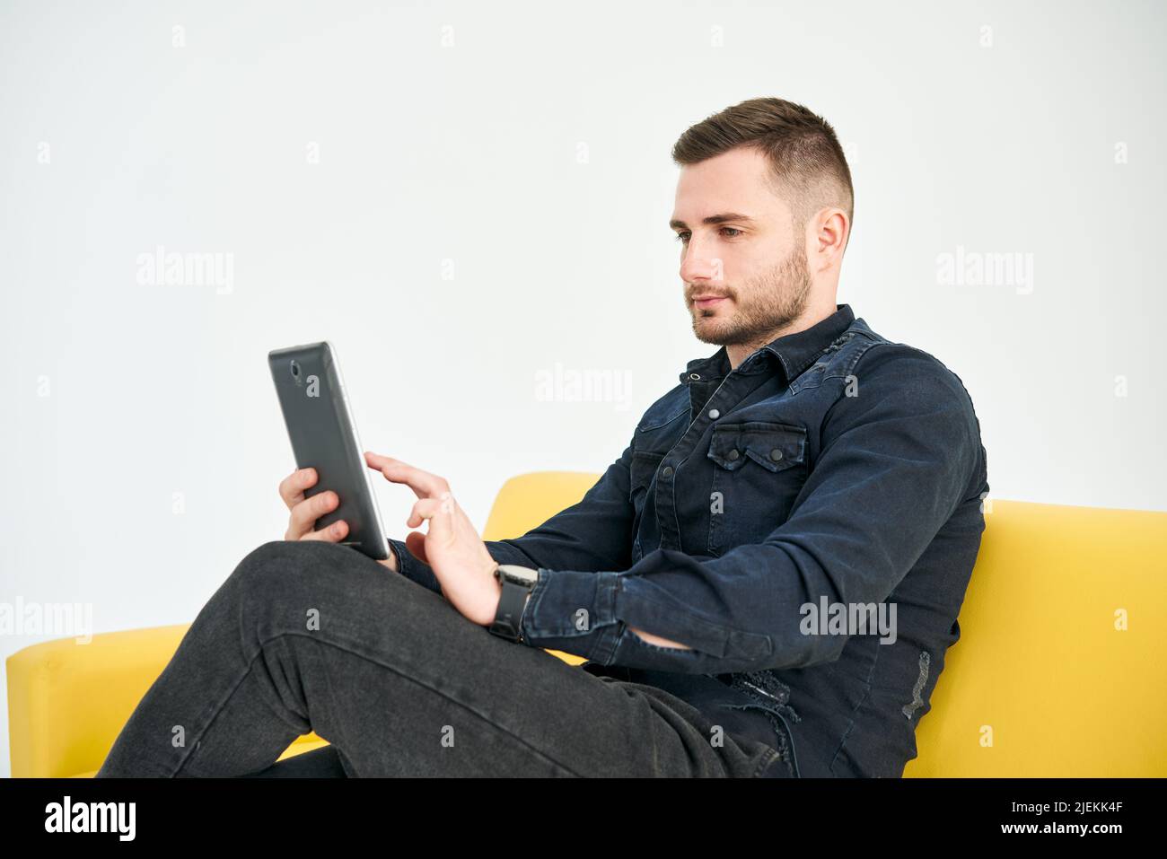 Giovane uomo che guarda il tablet digitale mentre si siede su un divano luminoso. Business, freelance, concetto digitale Foto Stock