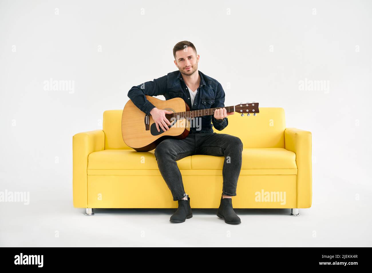 Bel giovane uomo che suona la chitarra acustica che si siede su un divano giallo su sfondo bianco. Hobby, concetto di musica Foto Stock