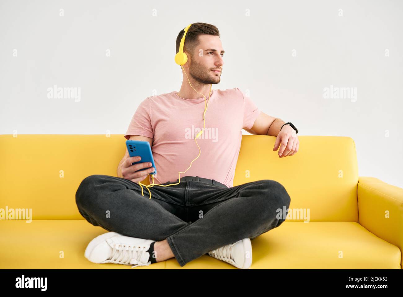 Il giovane uomo rilassato indossa le cuffie per ascoltare la musica dal suo telefono cellulare mentre si siede sul divano giallo su sfondo bianco Foto Stock