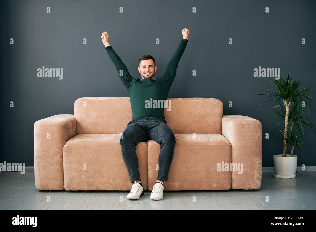 Giovane gioioso con le braccia alzate celebrando il successo seduto sul divano in appartamento moderno. Concetto del vincitore Foto Stock