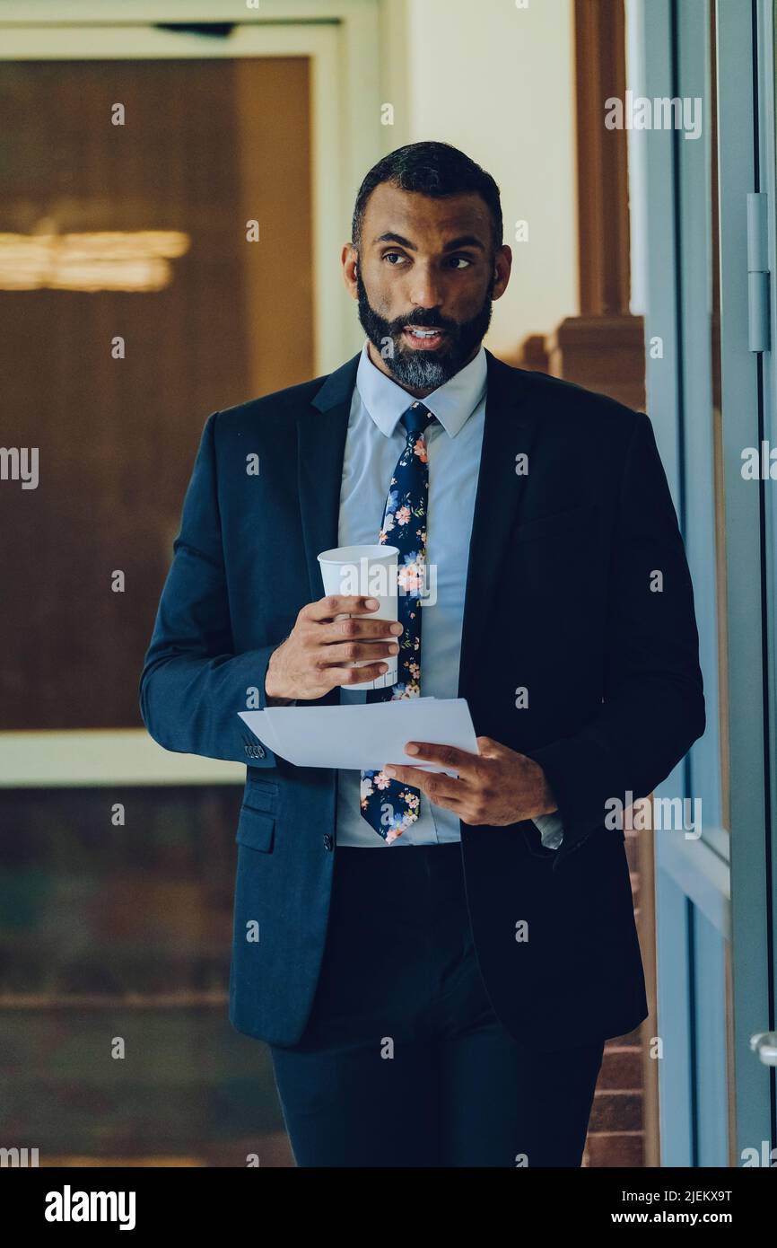 MID adulto bearded uomo nero imprenditore uomo d'affari che indossa il vestito che tiene la tazza di caffè e le carte che camminano in ufficio colpo Foto Stock