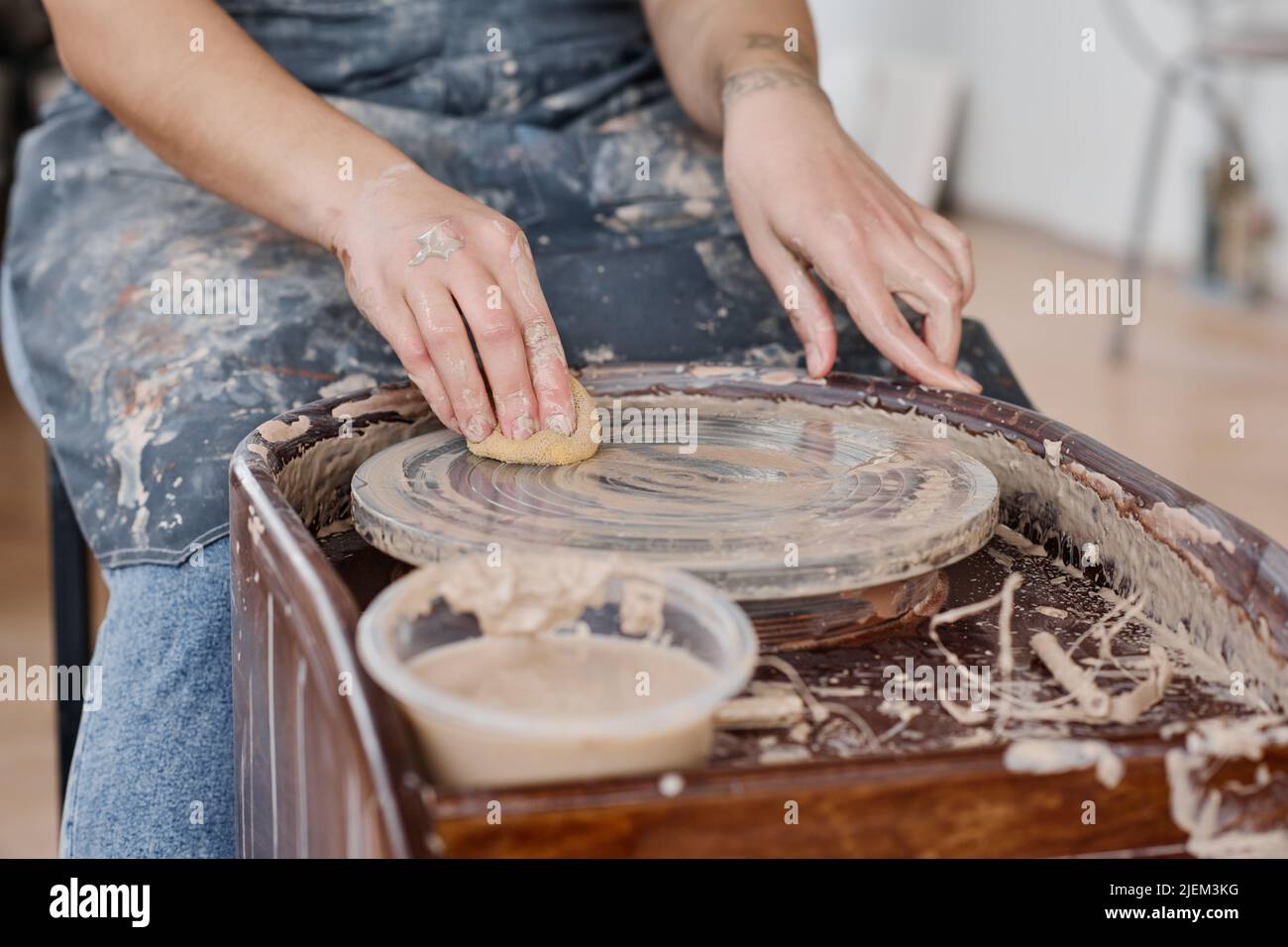Mani di giovane donna che puliscono la ruota di ceramica con spugna bagnata dopo la creazione di nuovi articoli di terracotta per la vendita in officina o studio Foto Stock