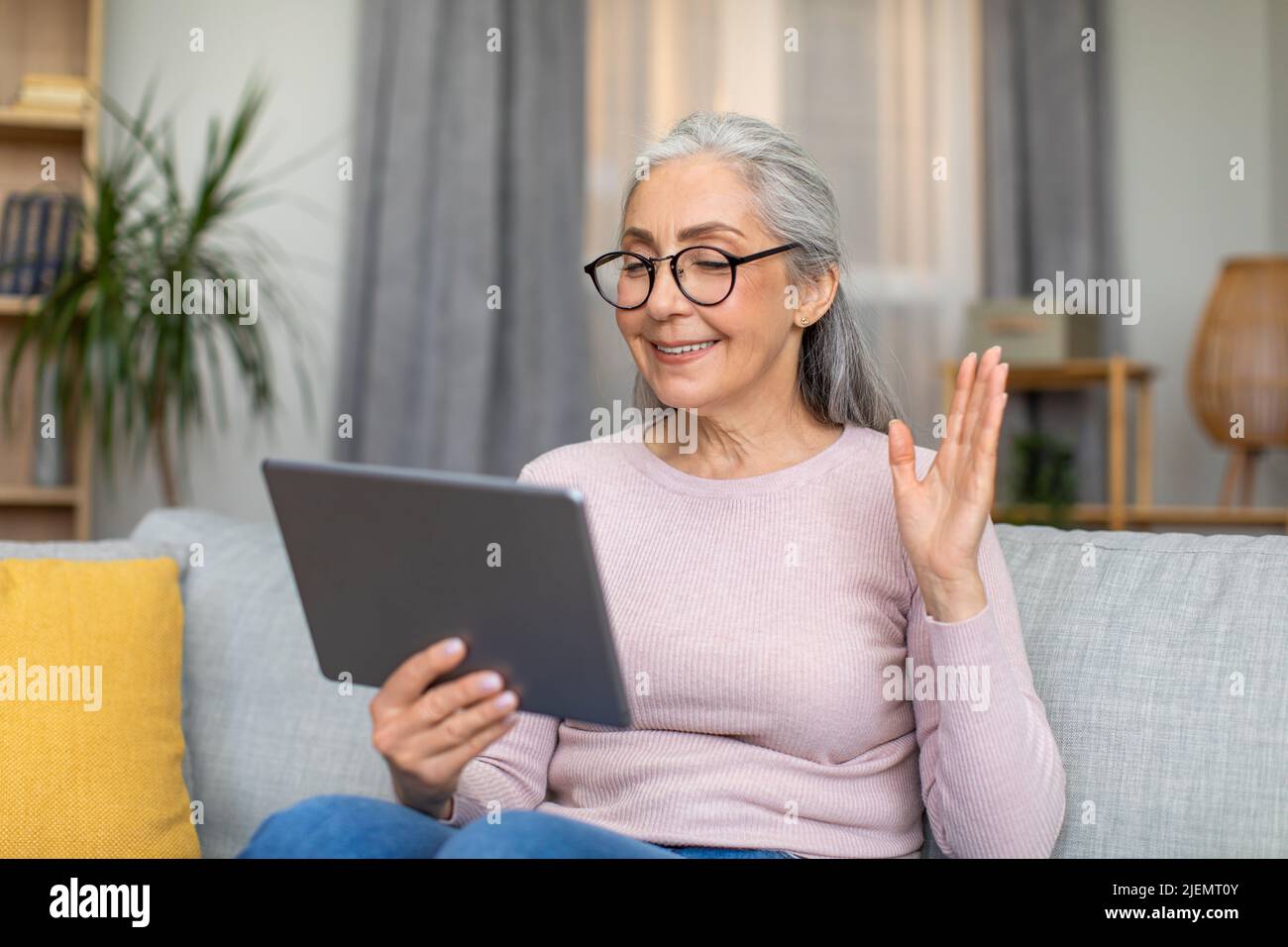 Felice europea matura donna con capelli grigi in occhiali onde la sua mano e guarda tablet, hanno riunione Foto Stock