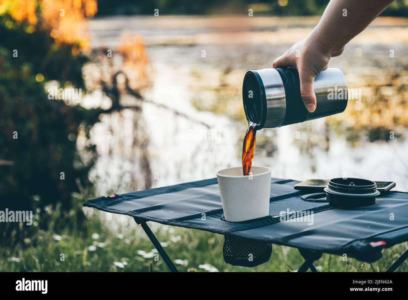 Versare il caffè nero caldo in una tazza di bambù riutilizzabile sul tavolo da campeggio all'aperto durante le prime ore del mattino. Preparazione di caffè appena fatto in natura Foto Stock