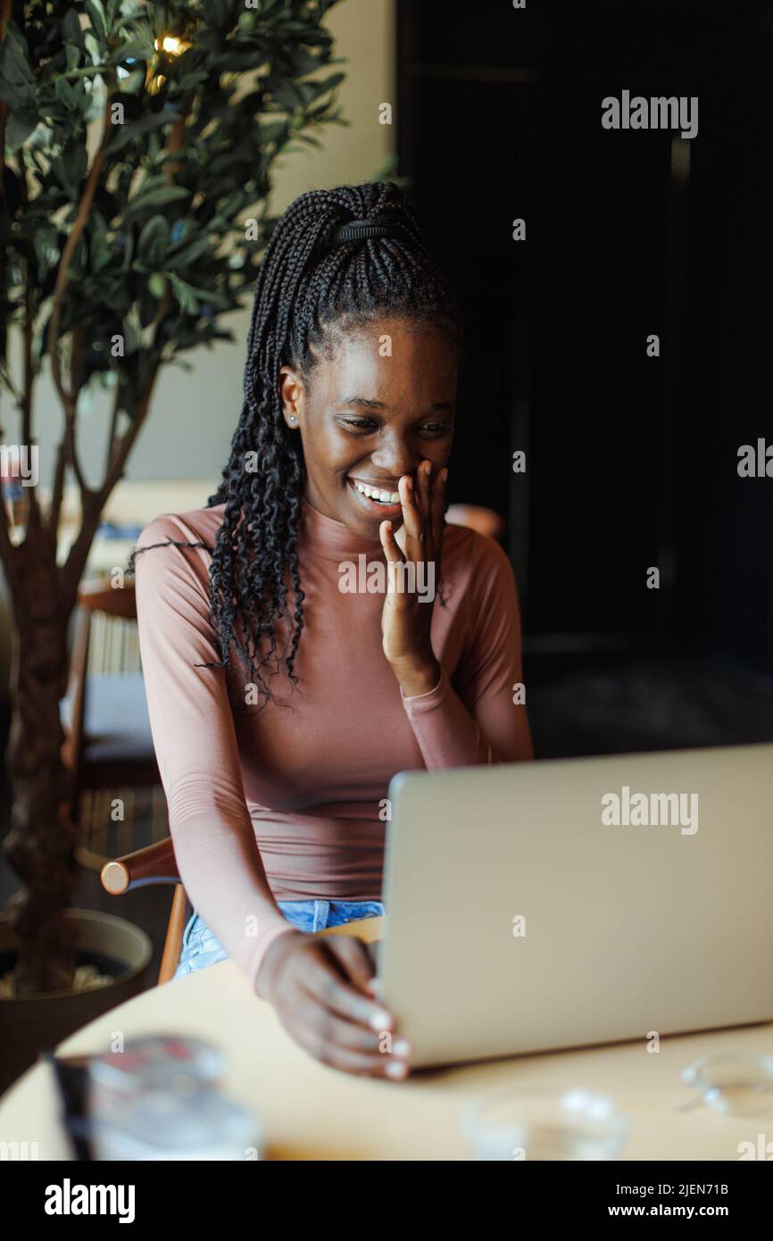 Ritratto di giovane donna afroamericana sorridente seduta al tavolo in un caffè, guardando il computer portatile, appoggiando la testa a portata di mano. Foto Stock