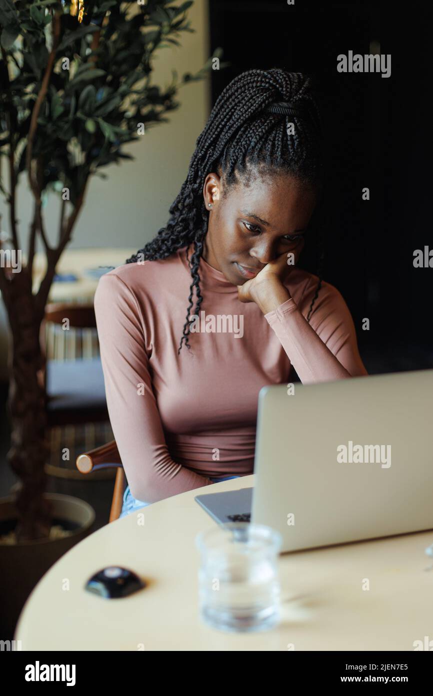 Ritratto di giovane donna afro-americana focalizzata seduta al tavolo beige in un caffè vicino a un computer portatile con le mani dietro la testa. Foto Stock