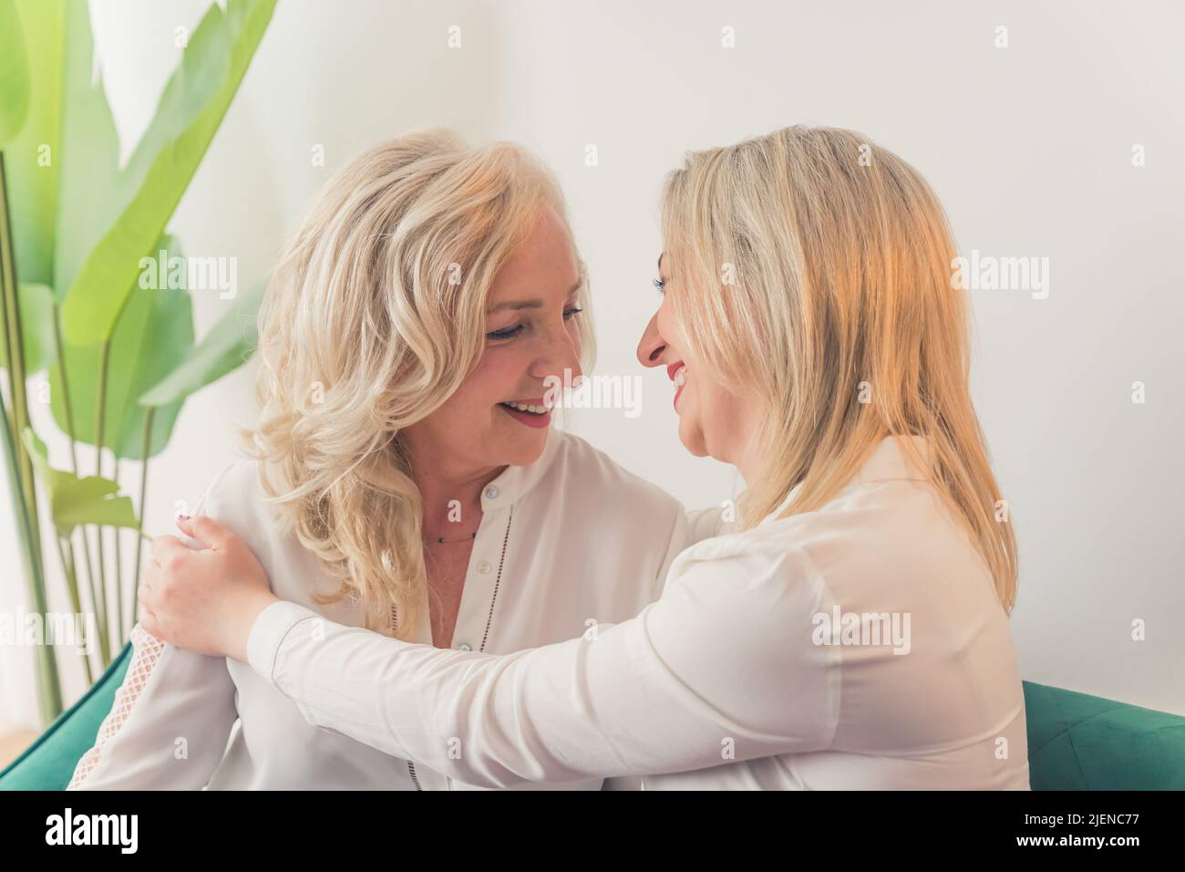 Primo piano ripresa interna di due donne caucasiche di mezza età con capelli biondi e camicie bianche seduti su un divano che si abbracciano e ridono. Foto di alta qualità Foto Stock