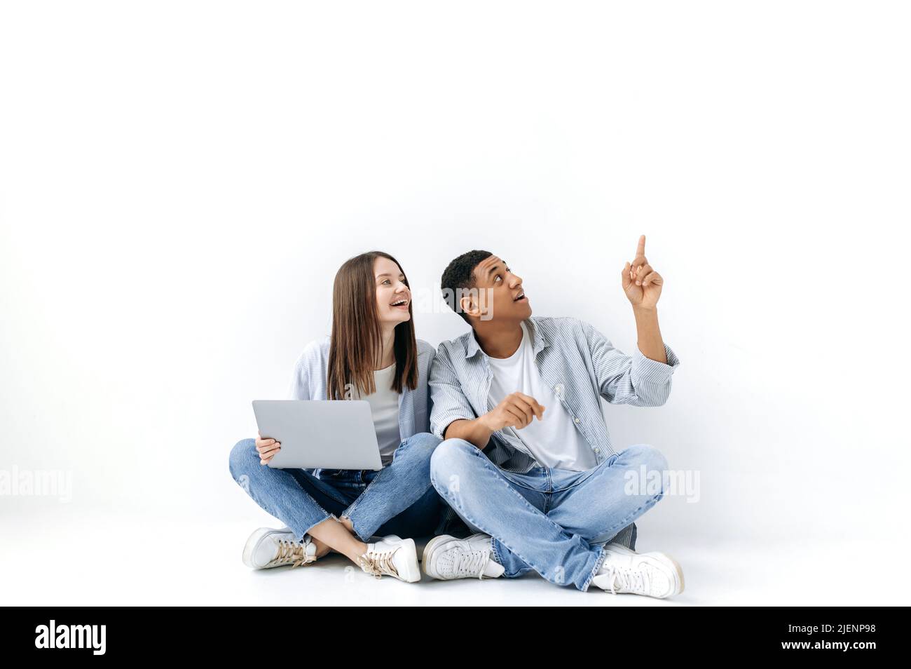 Bella donna caucasica e bell'uomo ispanico, coppia positiva, sono seduti con un computer portatile sul pavimento su sfondo bianco isolato, guardando lateralmente, il ragazzo punta con la mano a qualcosa Foto Stock