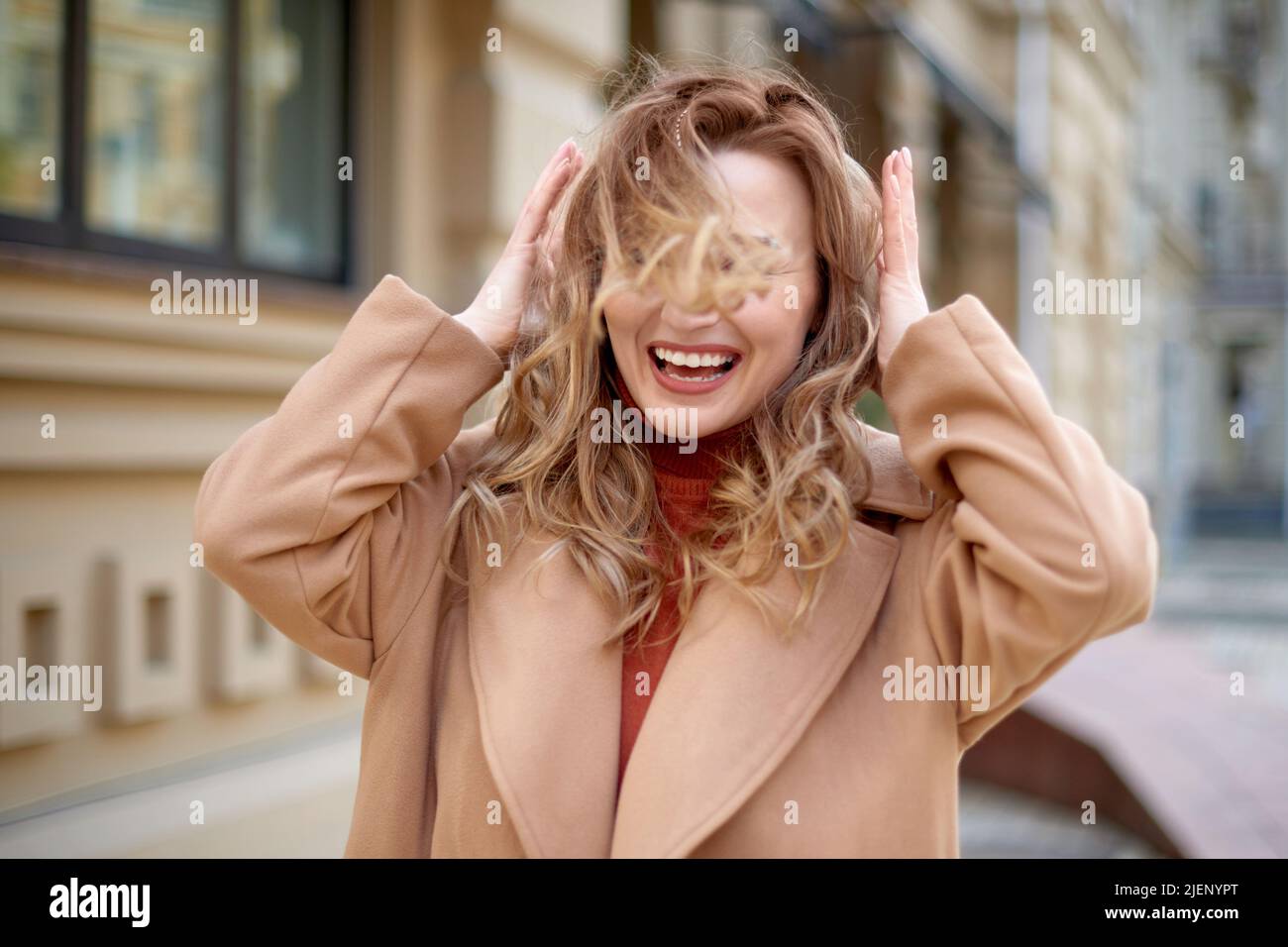 donna sorridente cammina al centro e protegge i capelli dal vento gusto in strada. le mani della ragazza tengono la testa e raddrizzano i capelli Foto Stock