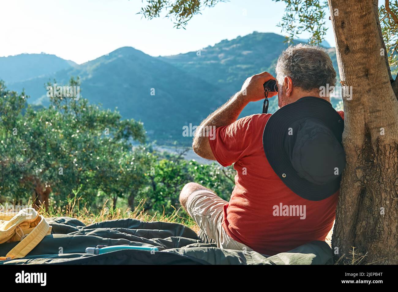 Uomo turistico riposato in oliveto, guardando attraverso binocoli alla vista delle montagne. Viaggiare o allenarsi all'aperto. Persone attive, escursioni, sano Foto Stock