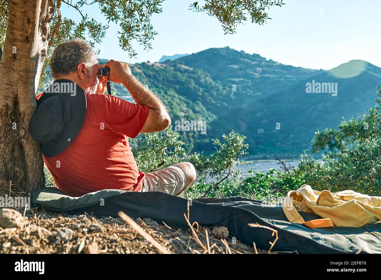 Uomo turistico riposato in oliveto, guardando attraverso binocoli alla vista delle montagne. Viaggiare o allenarsi all'aperto. Persone attive, escursioni, sano Foto Stock