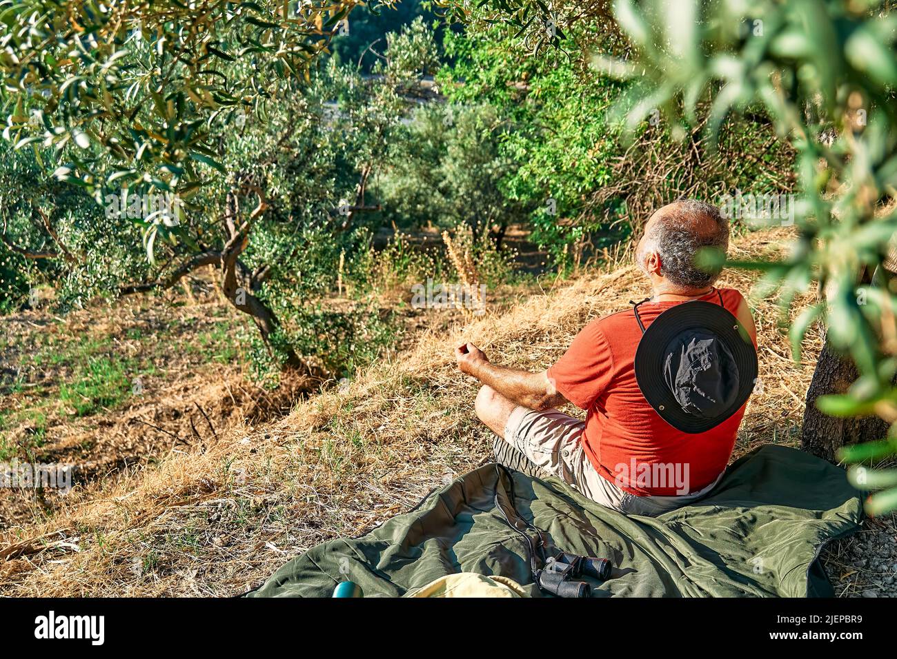 Uomo turistico che riposa in uliveto, meditando di fronte alla montagna o esercitandosi all'aperto. Persone attive, viaggi, escursioni, stile di vita sano a Foto Stock
