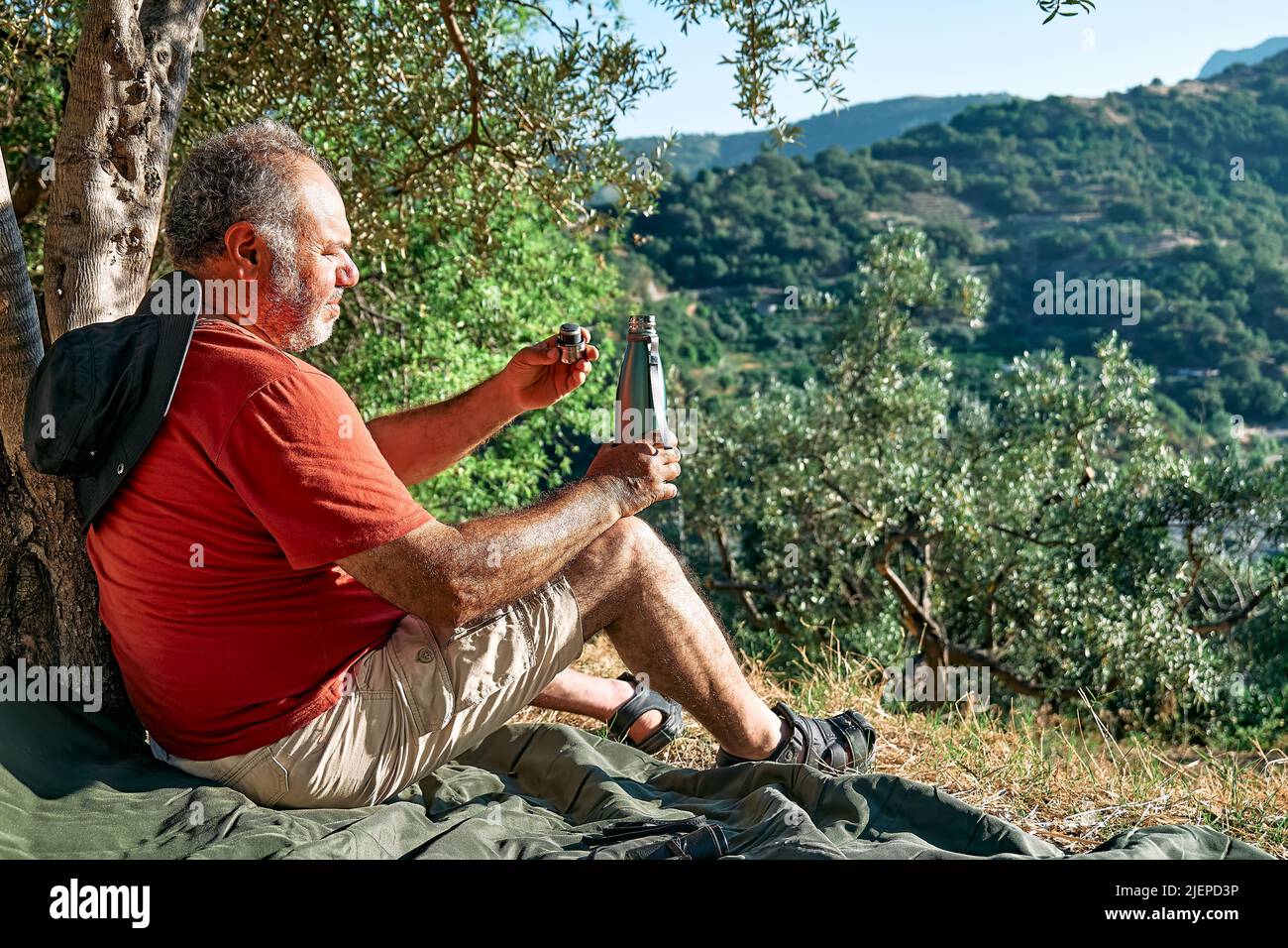 Uomo turistico che riposa in oliveto, bevendo acqua da bottiglia di acciaio termale, viaggiando o esercitandosi all'aperto. Persone attive, escursioni, vita sana Foto Stock