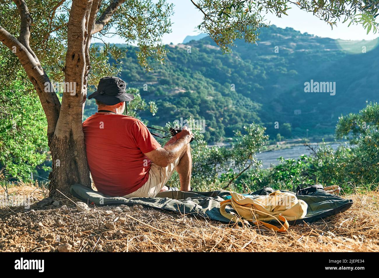 Uomo turistico che riposa in oliveto, godendo di vista montagna, viaggiando con zaino o esercitandosi all'aperto. Persone attive, escursioni, stile di vita sano a Foto Stock