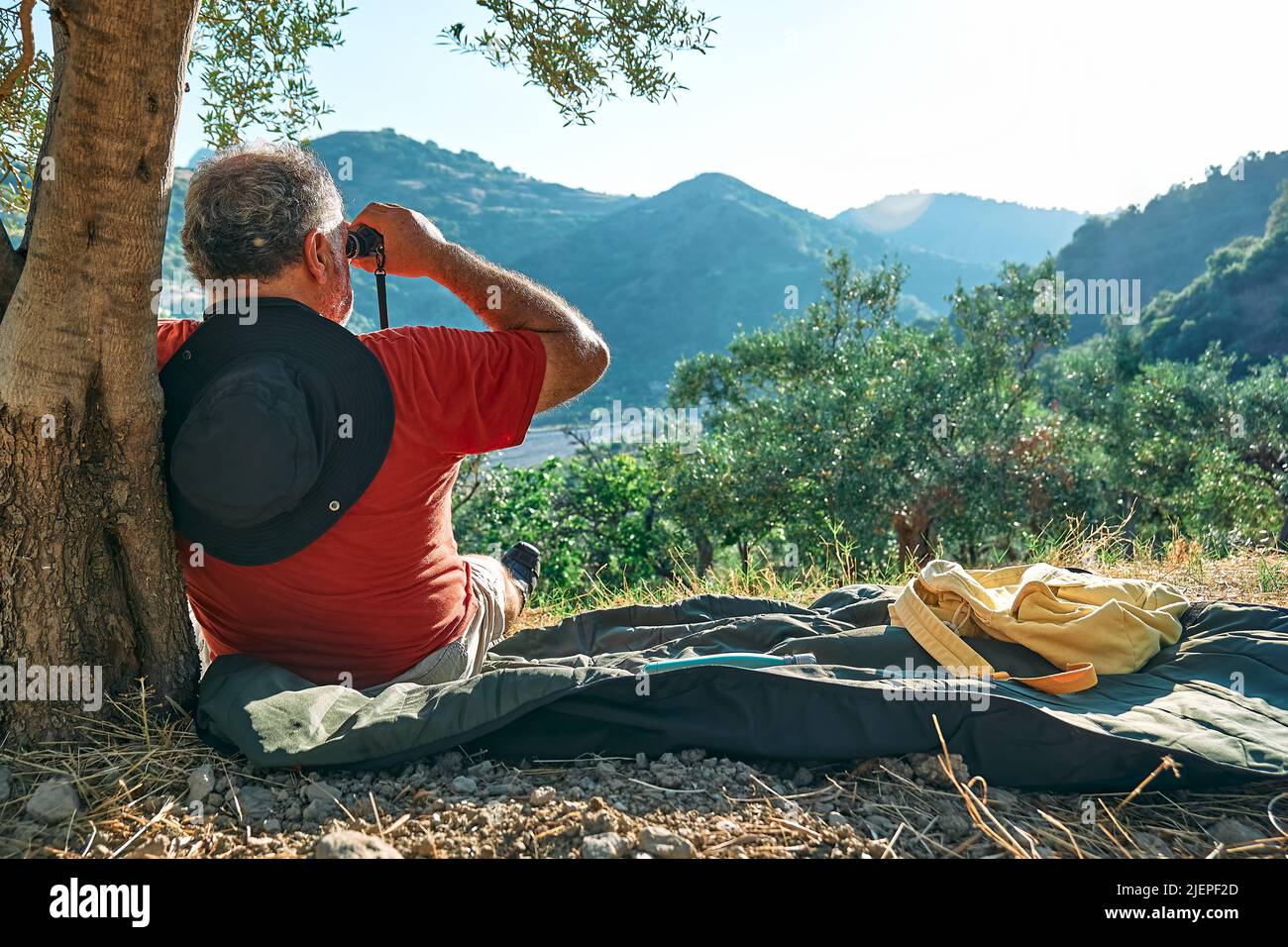 Uomo turistico riposato in oliveto, guardando attraverso binocoli alla vista delle montagne. Viaggiare o allenarsi all'aperto. Persone attive, escursioni, sano Foto Stock