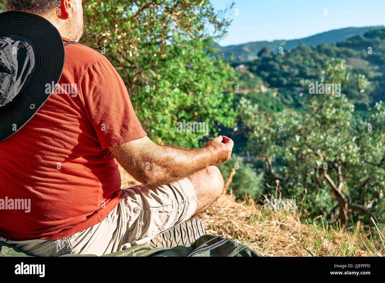 Uomo turistico che riposa in uliveto, meditando di fronte alla montagna o esercitandosi all'aperto. Persone attive, viaggi, escursioni, stile di vita sano a Foto Stock