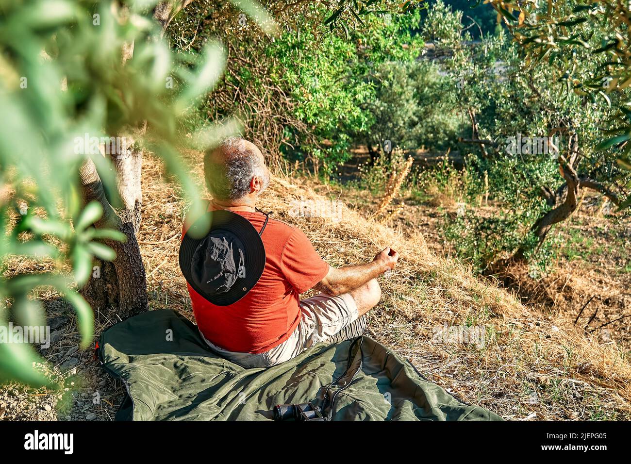 Uomo turistico che riposa in uliveto, meditando di fronte alla montagna o esercitandosi all'aperto. Persone attive, viaggi, escursioni, stile di vita sano a Foto Stock