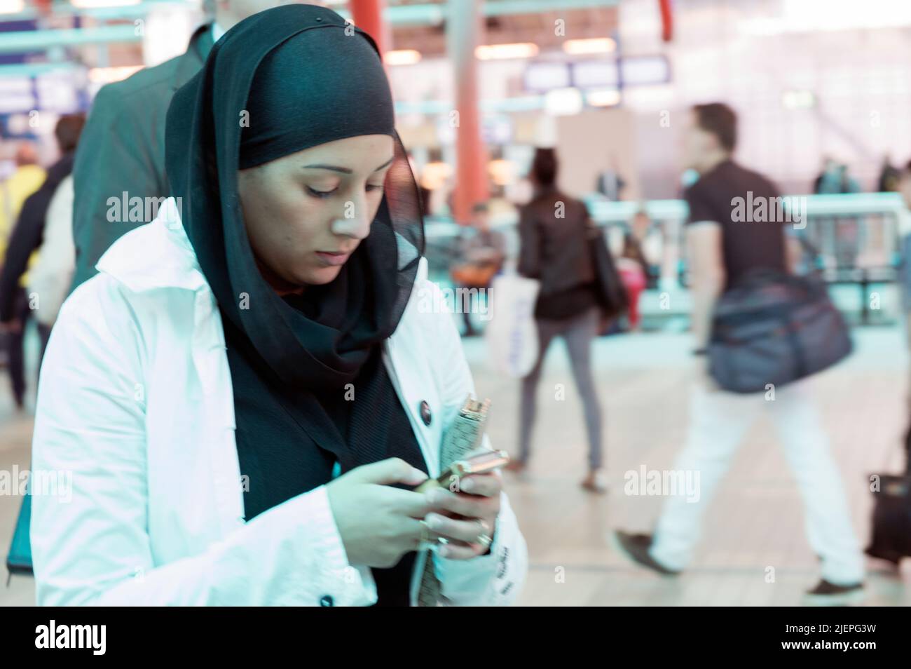 Utrecht, Paesi Bassi. Giovane donna araba adulta che indossa una sciarpa e il suo smartphone che cammina e cammina attraverso la stazione centrale mentre comunica, trasmette messaggi e naviga con il suo dispositivo smartphone. Foto Stock