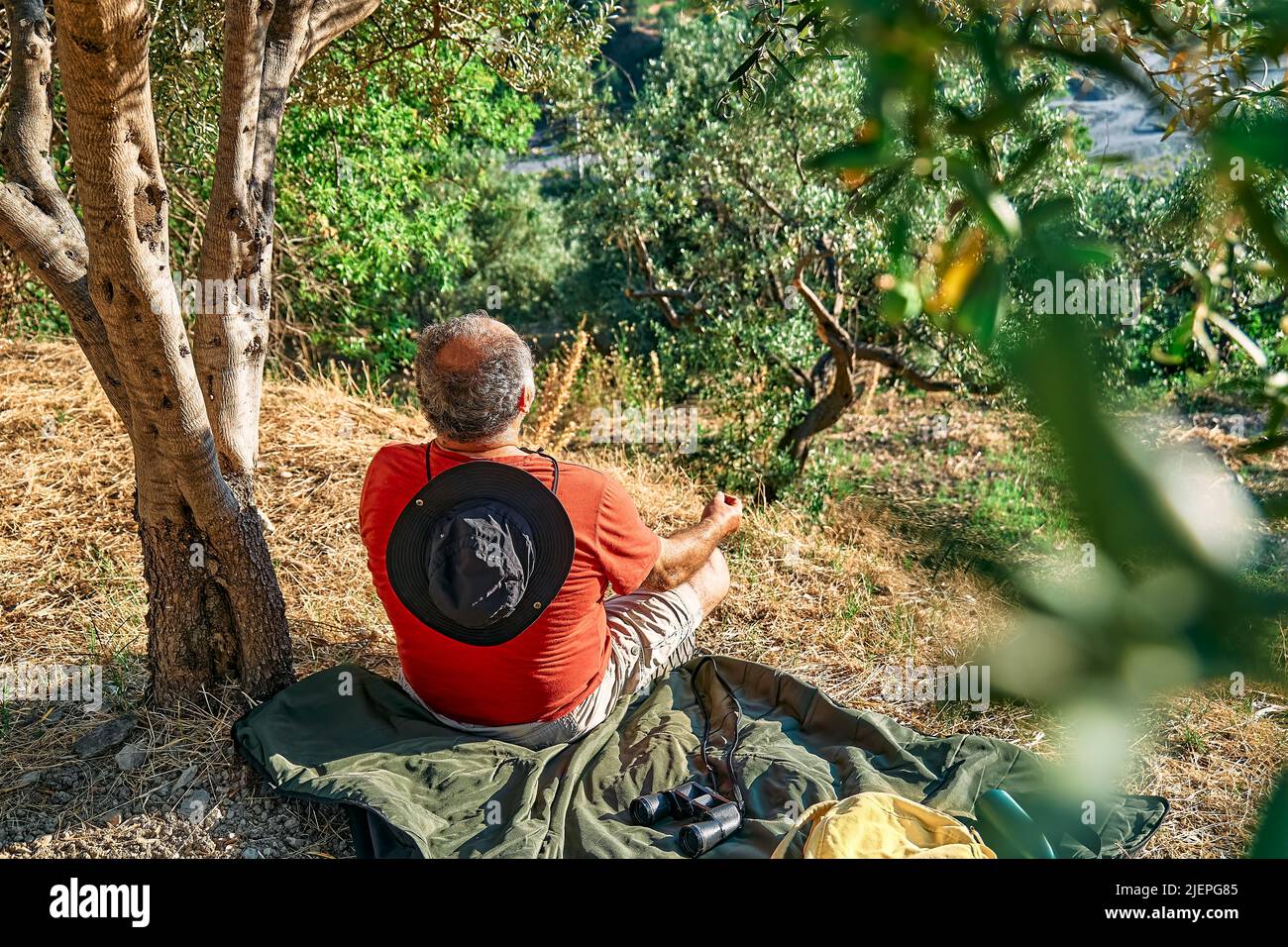 Uomo turistico che riposa in uliveto, meditando di fronte alla montagna o esercitandosi all'aperto. Persone attive, viaggi, escursioni, stile di vita sano a Foto Stock