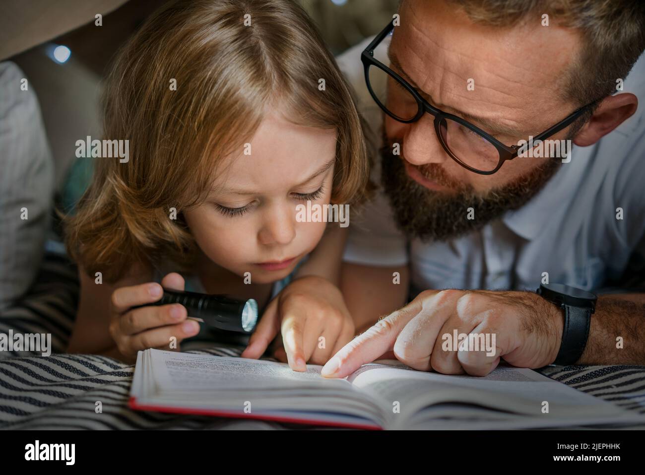 Ritratto di un ragazzo di 6 anni e suo padre che legge un libro in tenda teepee. Padre e figlio con libro di lettura torcia sotto coperta a casa. Foto Stock