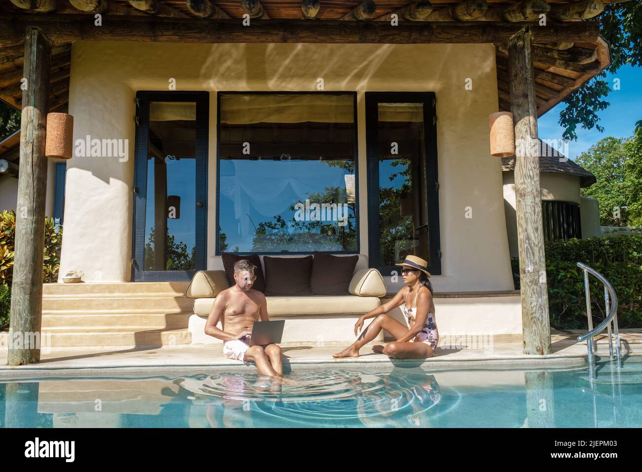 teleletto di lavoro digitale nomade uomini e donne e laptop seduta in una soleggiata piscina d'acqua turchese in una villa piscina privata, uomo d'affari usando mockup Foto Stock