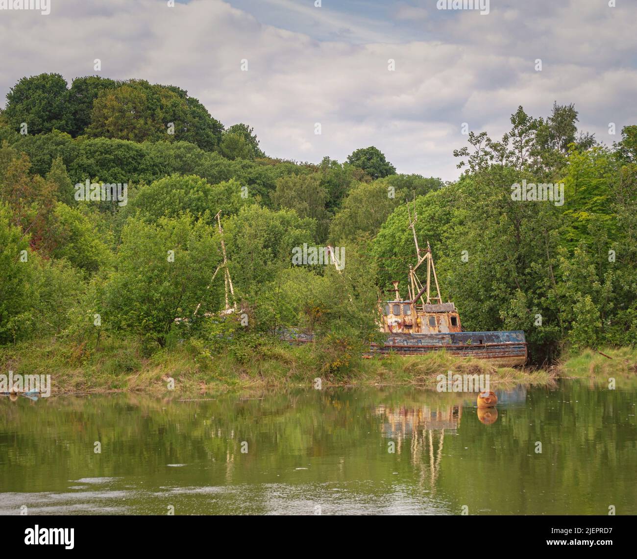 Una barca dilapidata e abbandonata si trova nascosta tra alcuni alberi. Lo scafo e la cabina sono arrugginiti e la vernice è incrinata. Foto Stock