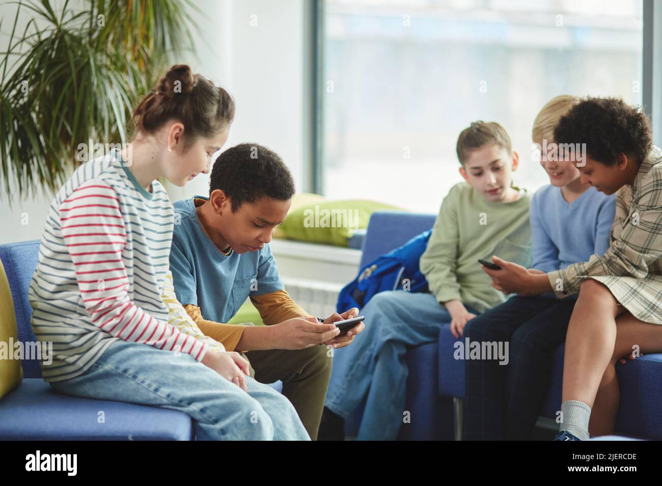 Gruppo di bambini diversi che usano gli smartphone a scuola durante la pausa, tutti vestiti con abiti colorati Foto Stock