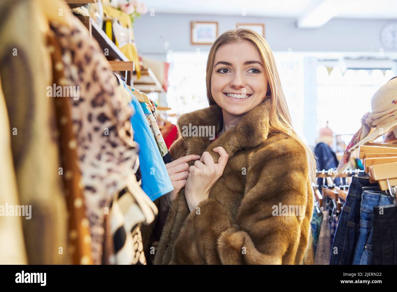 Giovane donna che acquista usato vestiti sostenibili dal negozio di beneficenza di seconda mano che prova su cappello Foto Stock