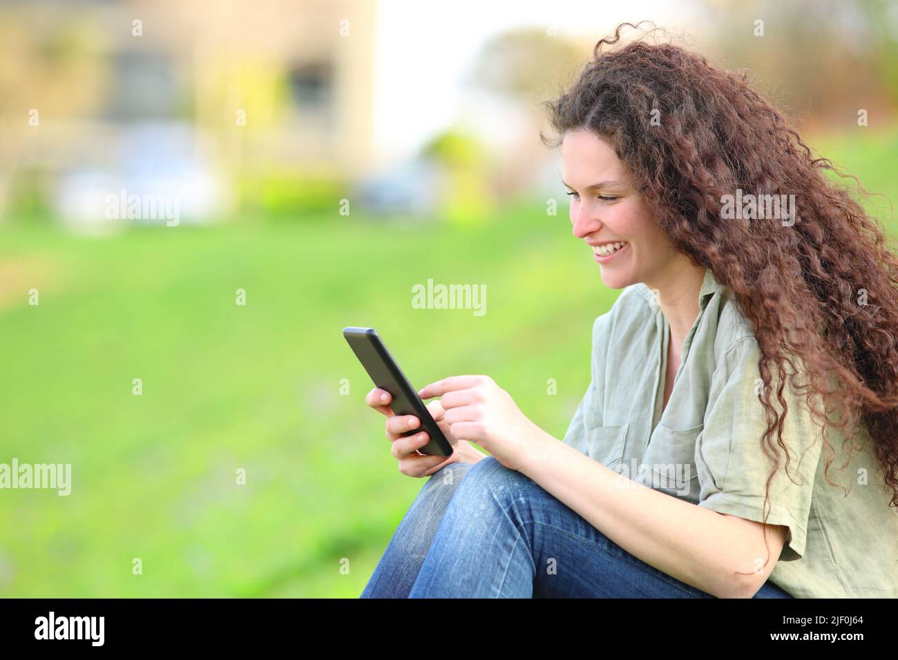 Donna felice con capelli ricci seduti utilizzando smartphone sul prato in un parco Foto Stock