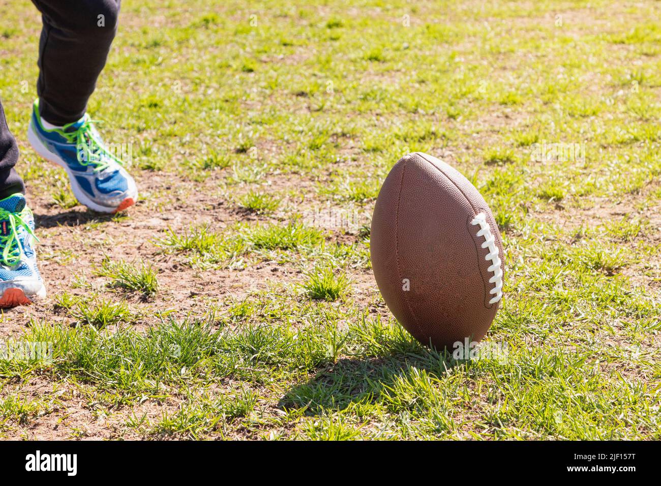 Su un campo d'erba c'è una palla da calcio americana e i piedi di una persona irriconoscibile indossando scarpe da ginnastica. Foto Stock