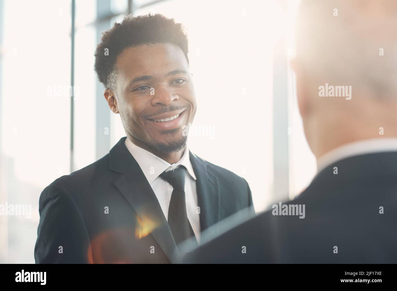 Positivo sicuro giovane uomo d'affari nero in tuta in piedi alla luce del sole e sorridendo al collega di affari mentre discutono il lavoro Foto Stock