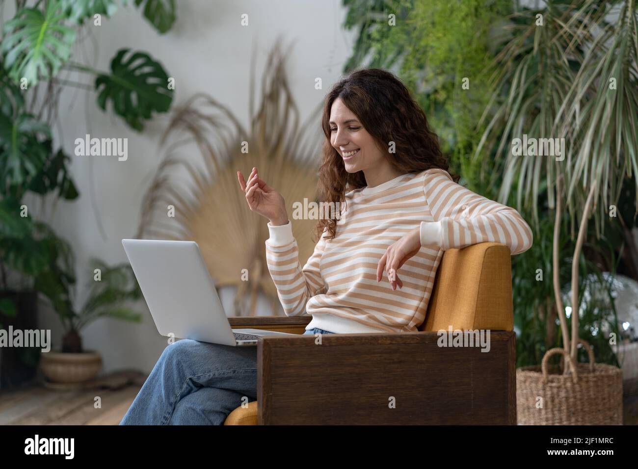 Donna italiana che sventola ciao, videochiamata su un portatile seduto in poltrona in un accogliente giardino di casa Foto Stock