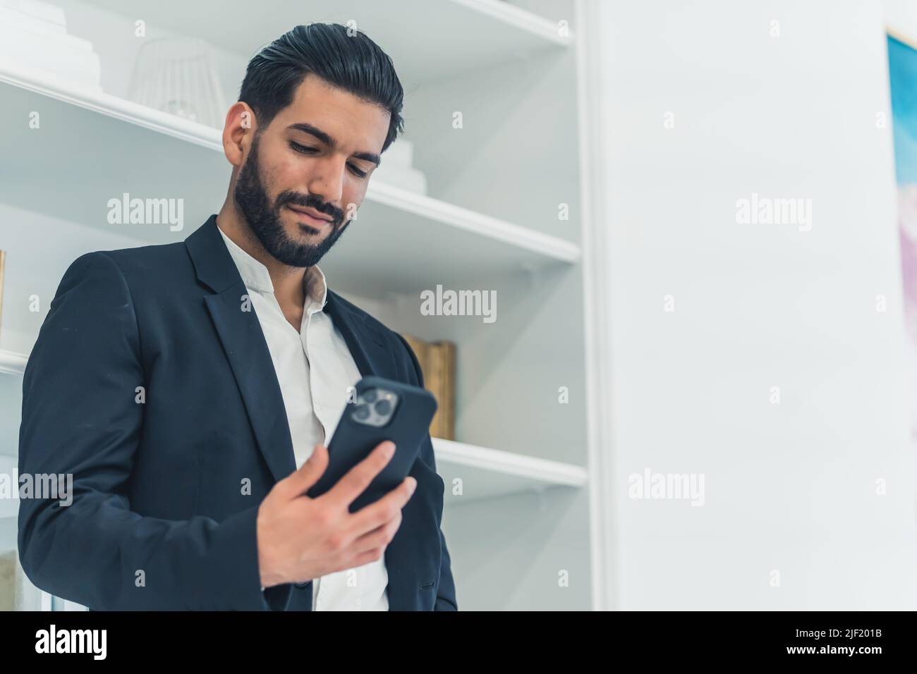 Soddisfatto felice buon-osservante unshaved uomo d'affari latino vestito in vestiti eleganti guardando il suo smartphone moderno. Scatto al coperto. Foto di alta qualità Foto Stock