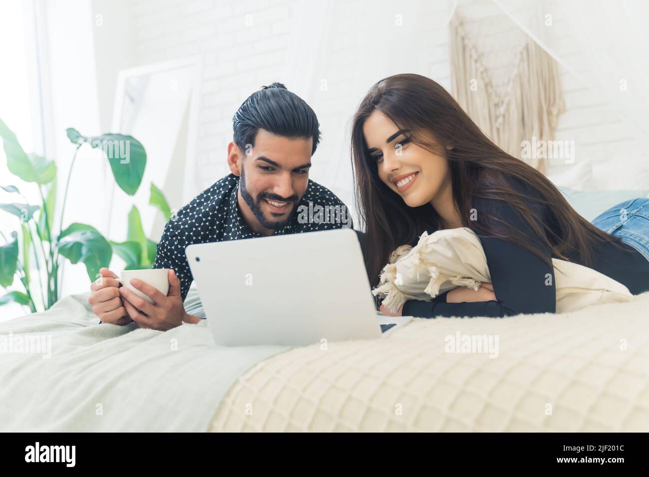 Cubano bello uomo dai capelli scuri e bruna europea bella donna sdraiata insieme sul letto guardando qualcosa sul portatile. Foto di alta qualità Foto Stock