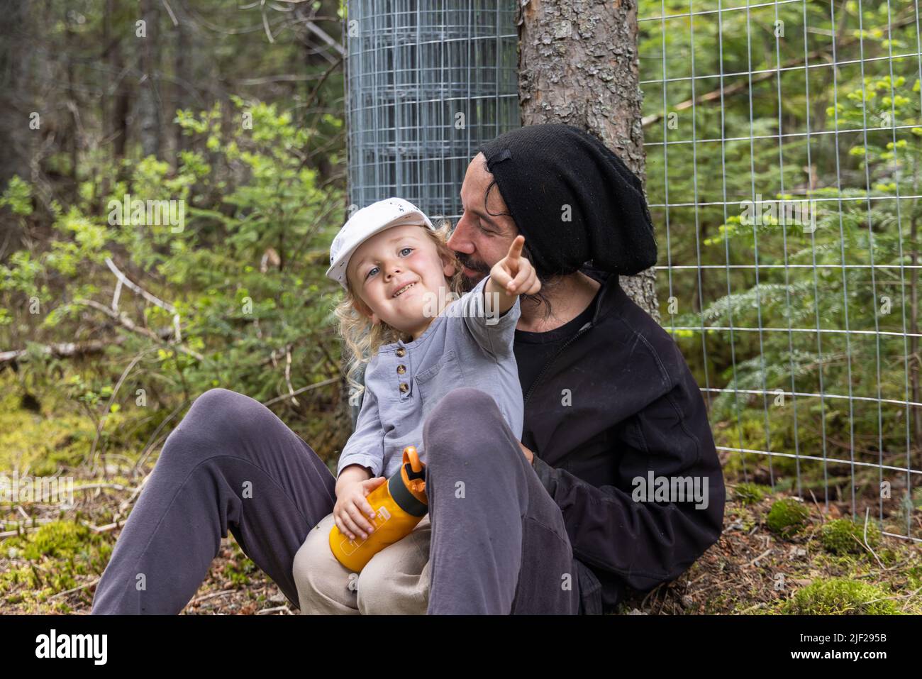 Felice padre e figlio giovane si riposano in natura. Ragazzo di due anni siede nel giro dei papà sorridendo e puntando il dito, con la foresta sfocata sullo sfondo. Foto Stock
